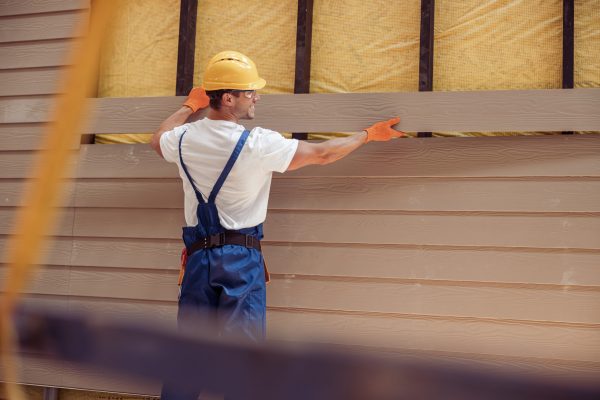 Cheerful young man builder wearing safety helmet and work overalls while installing exterior wood siding